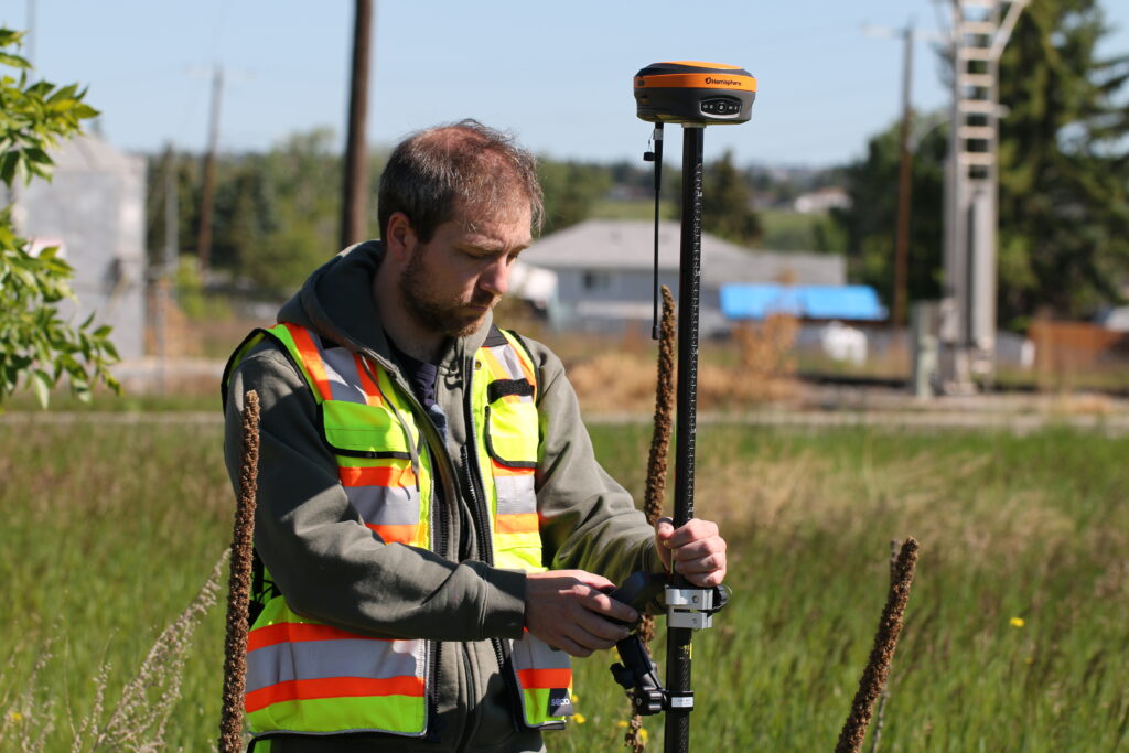 Surveyor in high-visibility gear operating a Hemisphere RTK GNSS rover near a residential utility corridor