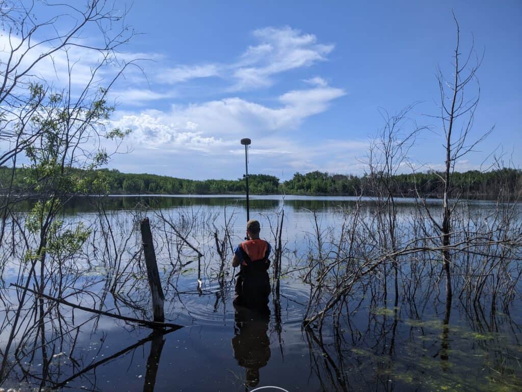Surveyor using aluminum GNSS surveying rod standing in shallow water during topographic survey