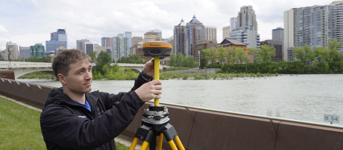 A man sets up a Hemisphere S631 GNSS Receiver in front of a city.